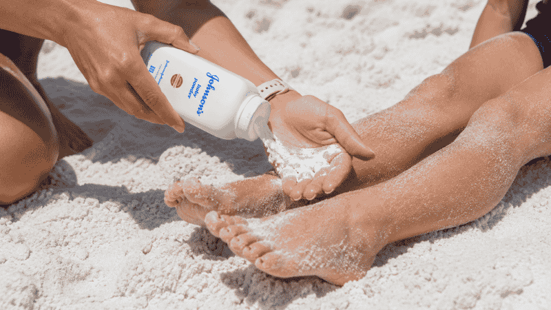 Person applying baby powder to sandy feet at the beach to remove sand easily