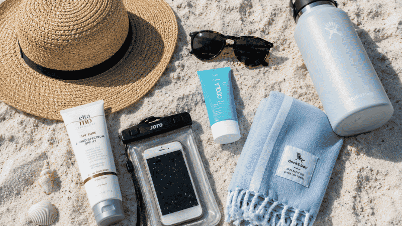 Flat lay of beach vacation packing list essentials including sunscreen, hat, water bottle and waterproof pouch on white sand