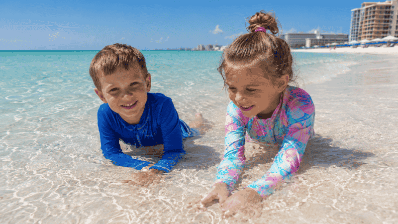 Children wearing UPF 50 rash guards playing in shallow Gulf water on a white sand beach