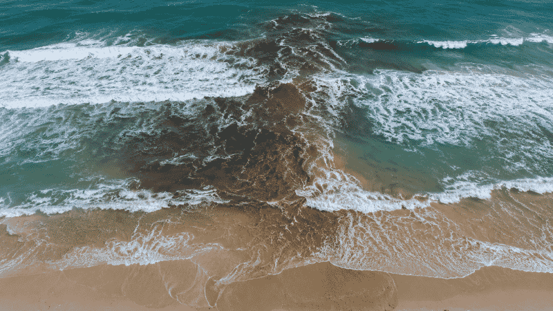 Aerial view of a rip current showing darker water cutting through breaking waves perpendicular to the beach shoreline