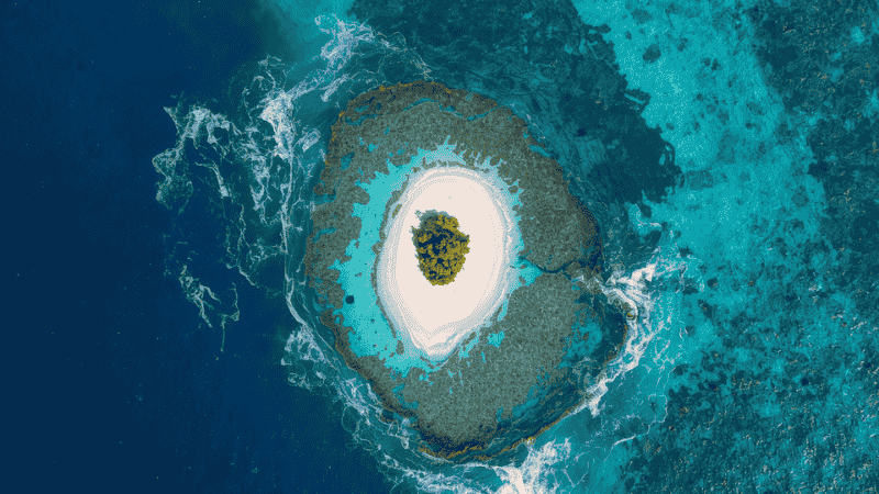 From a high altitude, looking down at the small island of Silk Caye in Belize, the white sandy beach is surrounded by huge, varying shades of blue coral reefs, presenting a typical all-inclusive island-hopping destination of Belize.