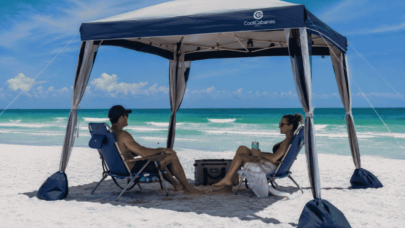 Open-sided beach cabana with sandbag legs on a windy white sand beach with couple relaxing inside
