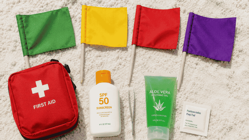 Beach safety essentials including colored warning flags first aid kit and sunscreen on white sand