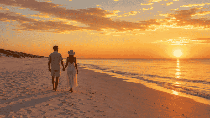 Couple walking on an empty white sand beach at sunrise with warm golden light reflecting on calm water