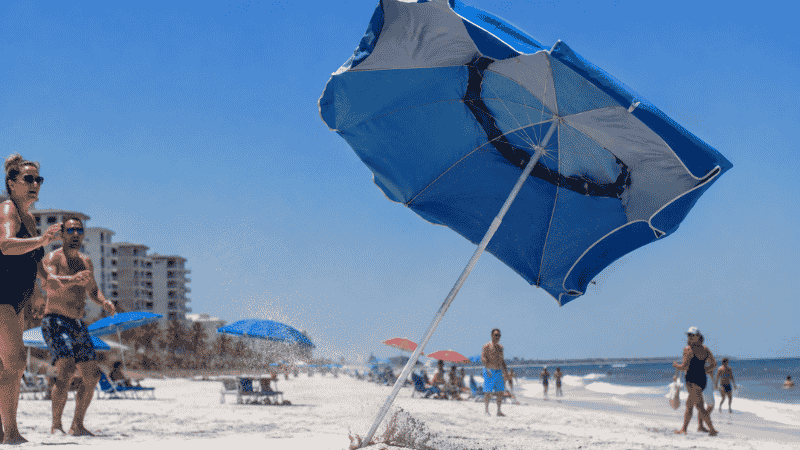Beach umbrella being blown sideways by strong coastal wind showing the safety hazard of improper anchoring