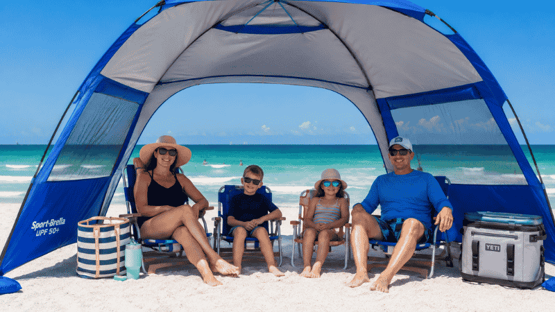 Family relaxing under a large pop-up beach tent on white sand Florida Gulf Coast beach with emerald water in background