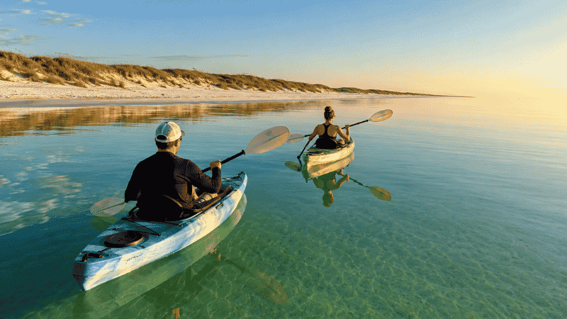 Two people kayaking on calm emerald water near a white sand Florida beach in early morning light