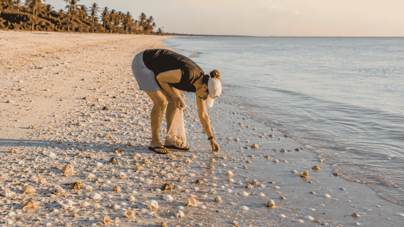 Person bent at the waist in the Sanibel Stoop position collecting seashells on white sand beach at Sanibel Island Florida