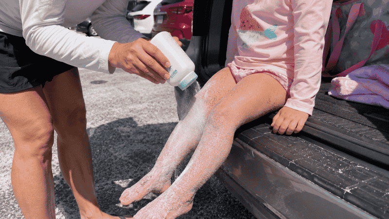 Parent applying baby powder to child's sandy legs at the beach to remove sand before getting in the car