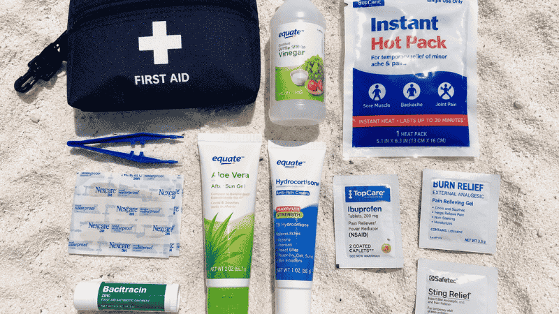 Beach first aid kit contents on white sand including tweezers vinegar heat pack waterproof bandages and aloe vera