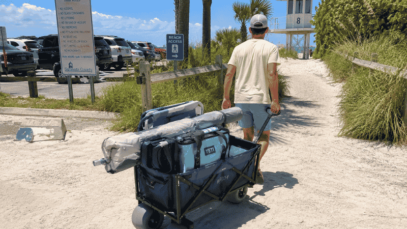 Person pulling a folding beach wagon with balloon tires through soft white sand loaded with beach gear