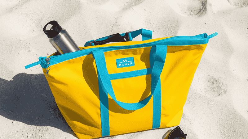 A brightly colored waterproof beach bag is placed on the white quartz sand. Inside the bag are zinc-based sunscreen, a stainless steel water bottle and polarized sunglasses.