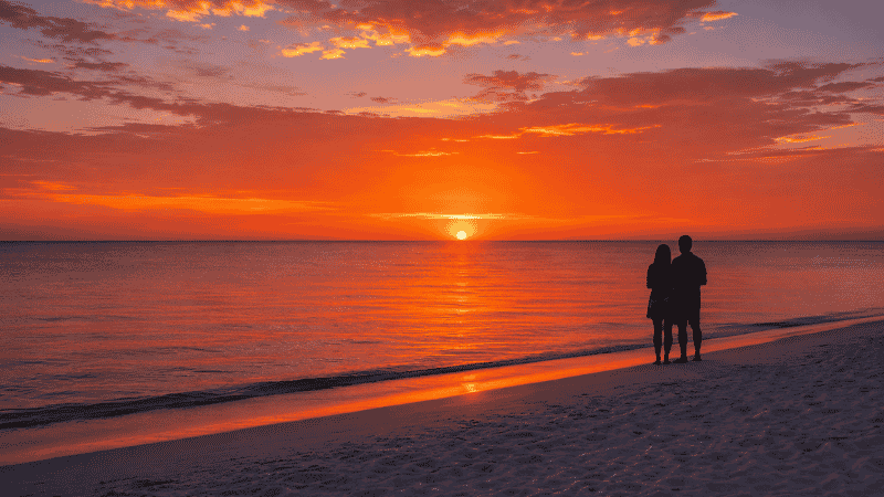 Perfect Gulf Coast Florida beach sunset with sun dropping into calm Gulf water and couple silhouetted on white sand beach