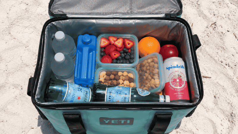Open insulated cooler bag on white sand beach showing organized food and drinks with ice packs
