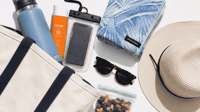 Minimal beach bag packed with seven essential items including sunscreen water bottle waterproof pouch towel hat and sunglasses