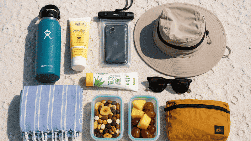 Complete beach gear essentials flat lay on white sand including sunscreen waterproof pouch hat sunglasses and water bottle