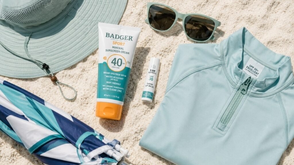 Flat lay of complete reef safe sun protection kit including mineral sunscreen UPF rash guard wide brim hat UV sunglasses SPF lip balm and beach umbrella on sandy beach background